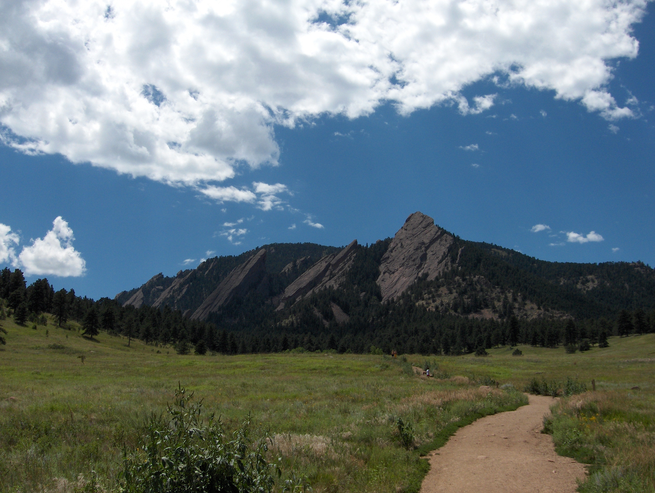 The Flatirons rise above a green meadow and trail in Boulder, Colorado